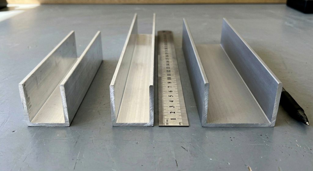 Three silver-colored aluminium U-channel extrusions of varying dimensions resting flat on a plain grey workspace. The channels vary in width and wall height, showing the U-shaped cross-section facing upwards. A metallic ruler and a black pen lie parallel to the extrusions on the right side to provide a visual scale. The lighting is bright, highlighting the brushed, slightly matte finish of the aluminium.