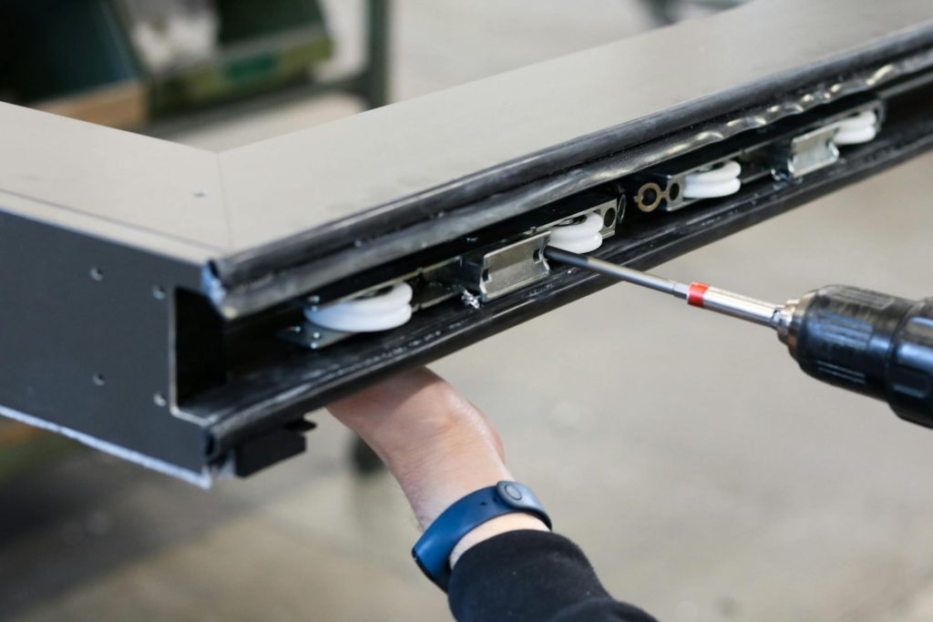A close-up of a black powder-coated aluminium extrusion being fitted with roller components using a power drill during a window or sliding door assembly.